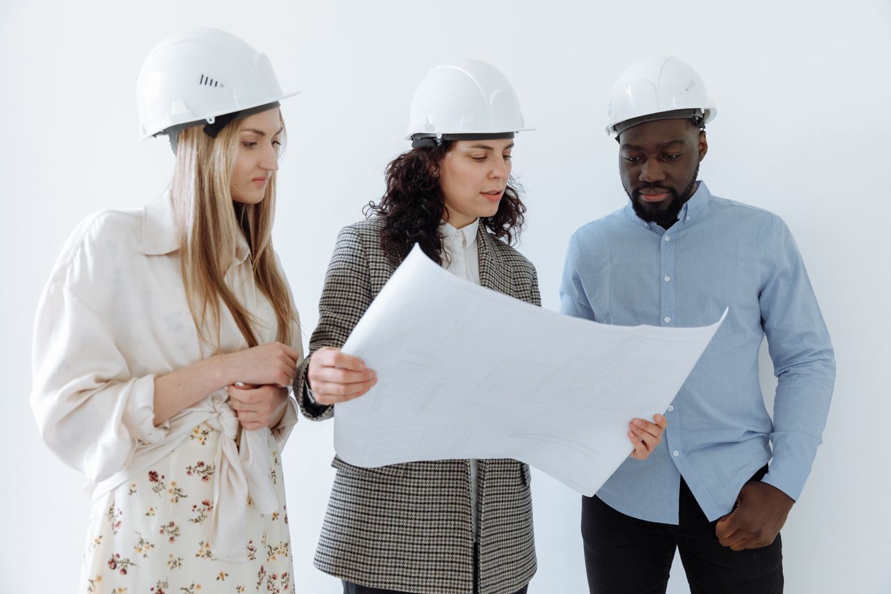 services-02 Three architects wearing hard hats reviewing blueprints indoors for a construction project.