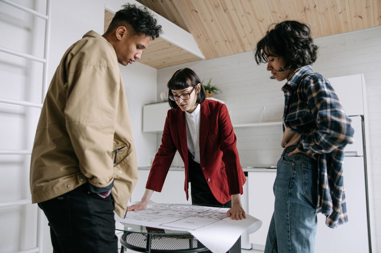 who-we-are A real estate agent discusses property plans with a young couple indoors.